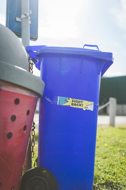 A tall, bright blue wheeled rubbish bin with a closed lid, situated on a paved or grassy outdoor area. The bin features a yellow and blue sticker with the message 'Fight Back!' adhered to its front su